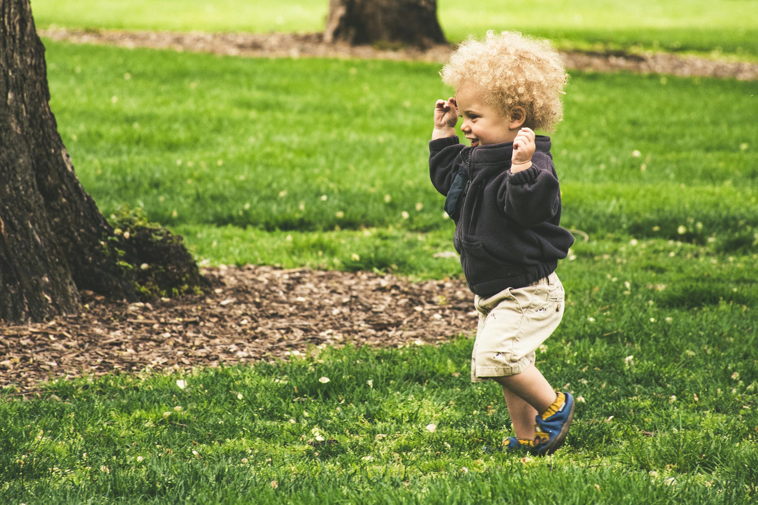 A happy toddler with curly hair joyfully runs on grass in a sunny park.