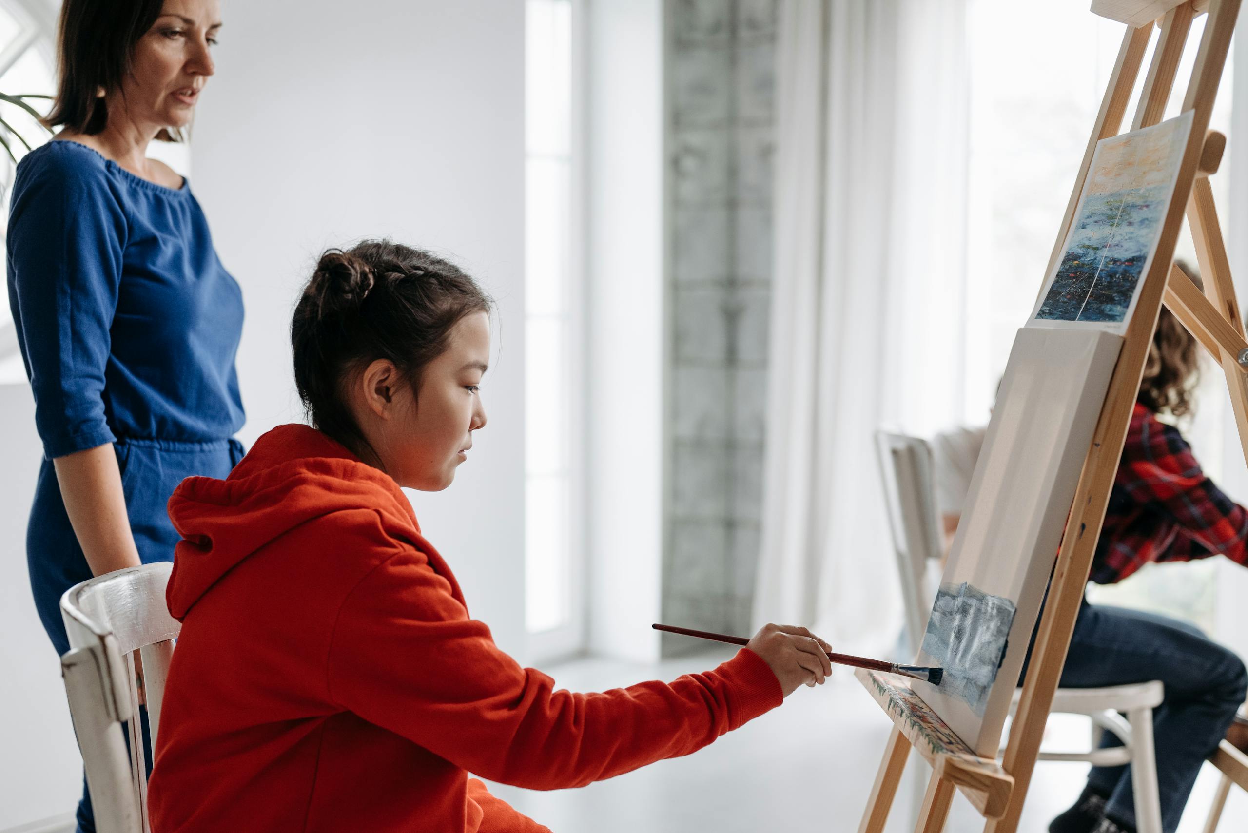 A young girl in a red hoodie painting on a canvas in an art class with teacher supervision.