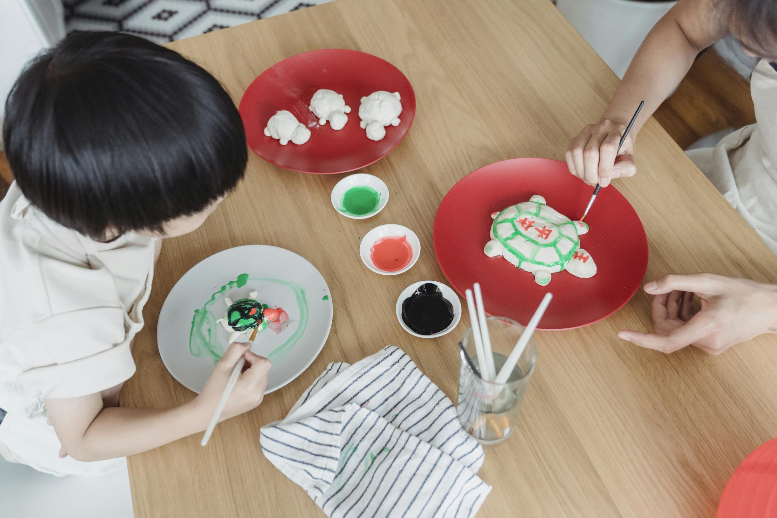 Child and parent decorating dough with paint at a wooden table, creating art together.