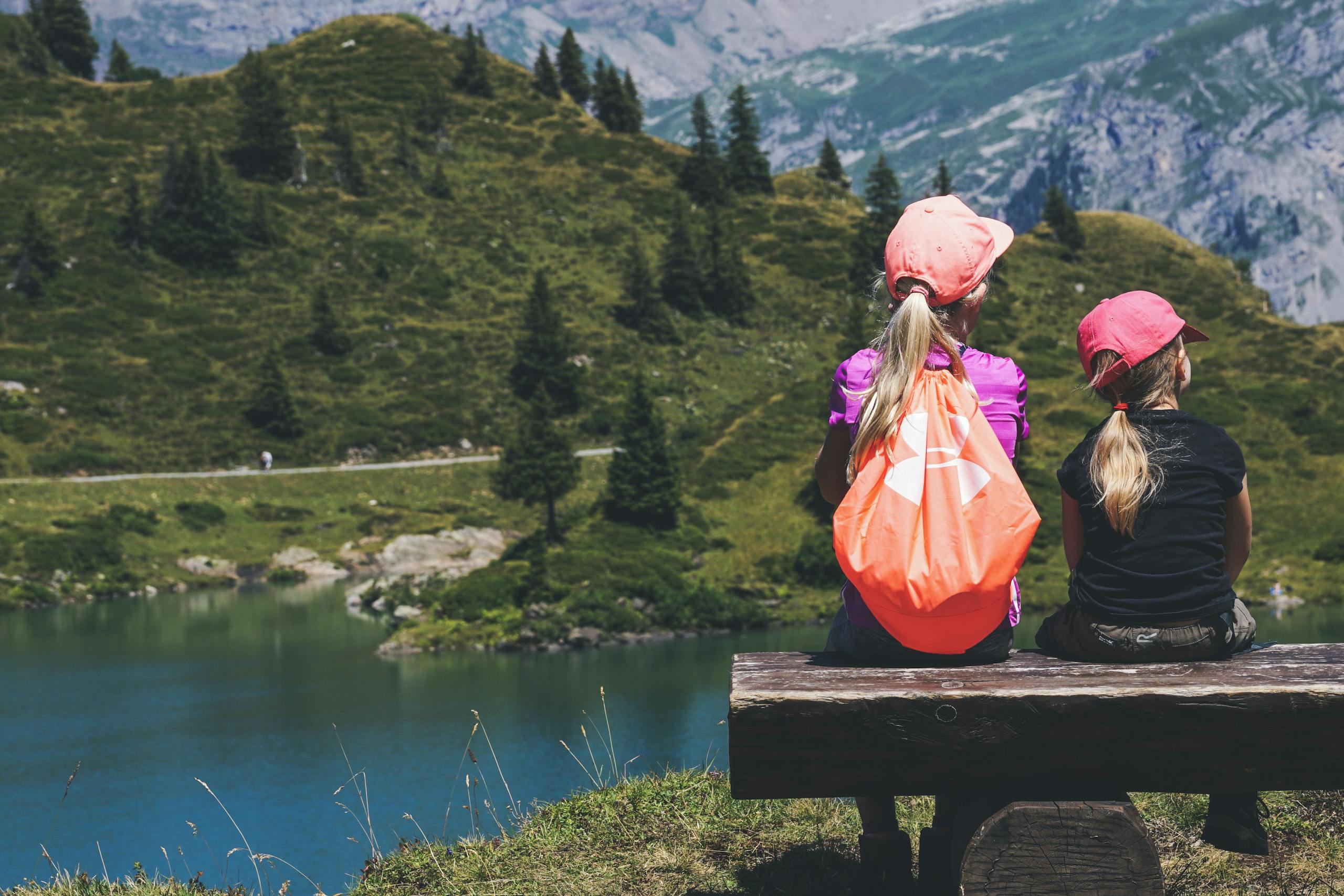 Two young girls sitting on a bench overlooking a serene mountain lake, enjoying a summer hike.